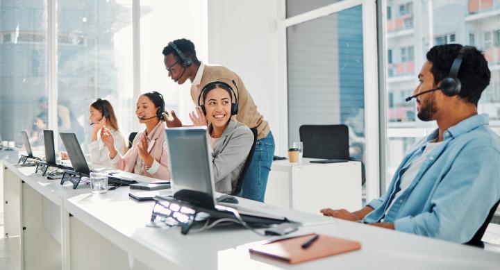 A diverse group of IT professionals sitting at long white table with computers. Each person has a headphones set. The room is brighly lit from a large window in the background.